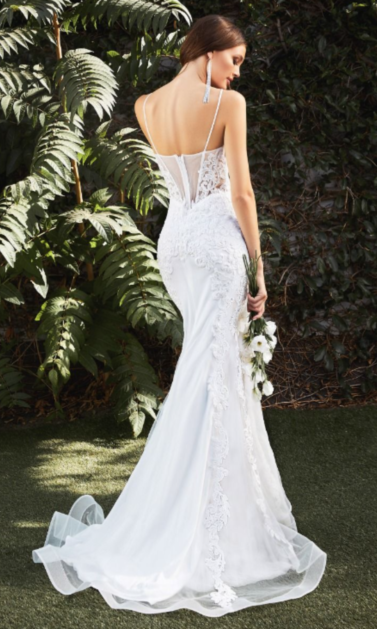 A woman in the Ladivine CD937W Long White Lace Formal Dress stands outdoors, holding white flowers and facing away from the camera.